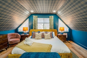 A bedroom with a sloped ceiling, a large bed with multiple pillows, two bedside lamps, a chair, and a window with curtains.