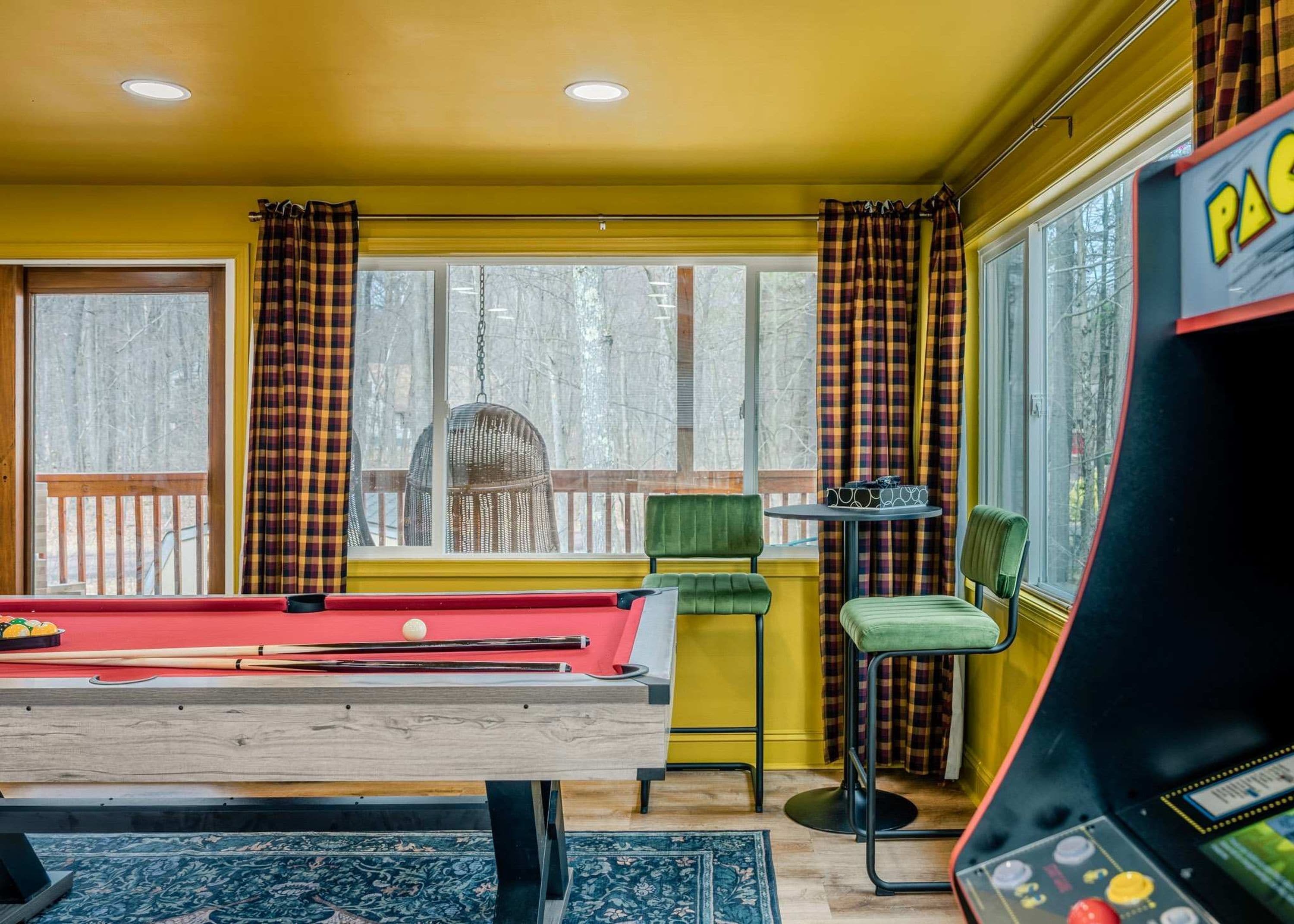 A room with a pool table, chairs, a table with glasses, and a view of trees outside.