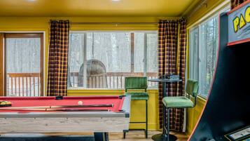 A room with a pool table, chairs, a table with glasses, and a view of trees outside.