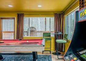 A room with a pool table, chairs, a table with glasses, and a view of trees outside.