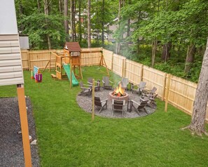 Un patio trasero con una casita de juegos de madera, un tobogán, un hogar para fogata con sillas y una cerca de madera.