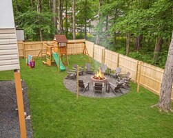 A backyard with a wooden playhouse, a slide, a fire pit with chairs, and a wooden fence.