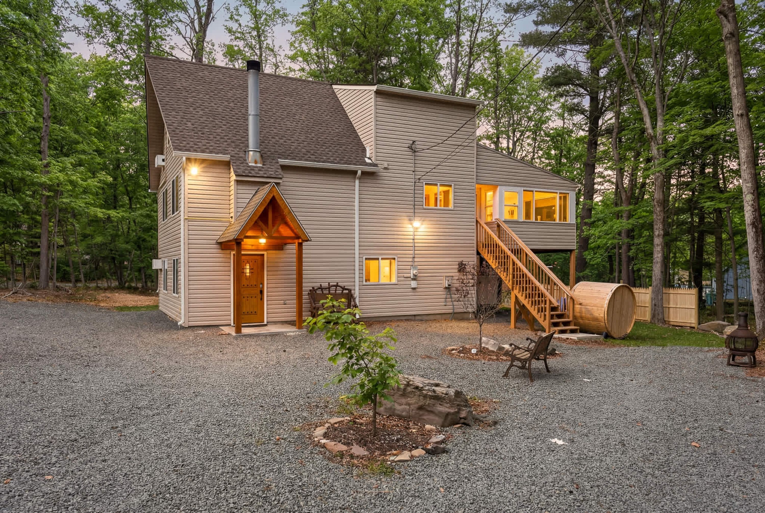 A two-story cabin with a gravel driveway, a wooden front door, a staircase, and a large barrel.