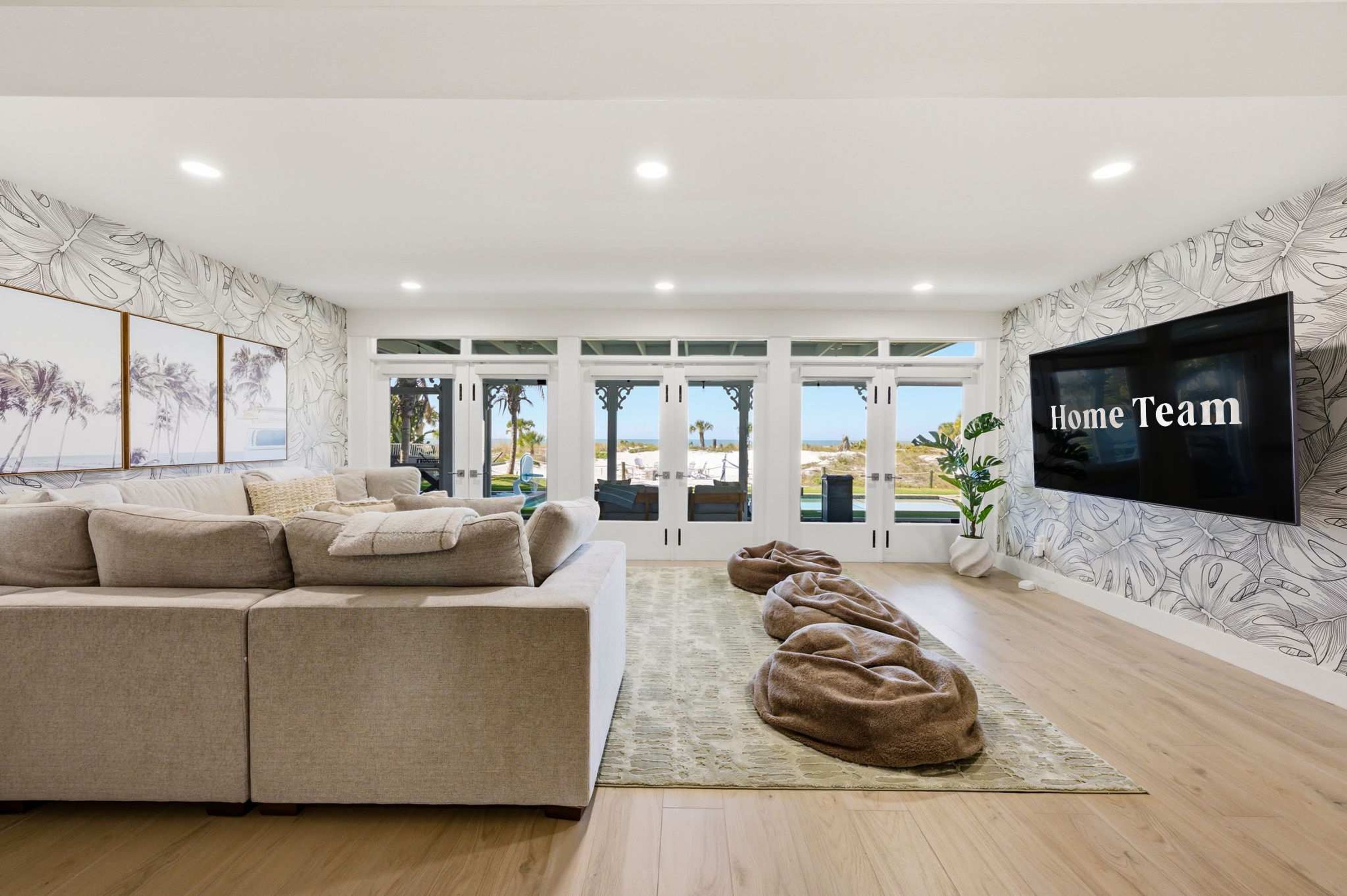 A modern living room with a large flat-screen TV, a beige sectional sofa, and a view of the outdoors through large windows.