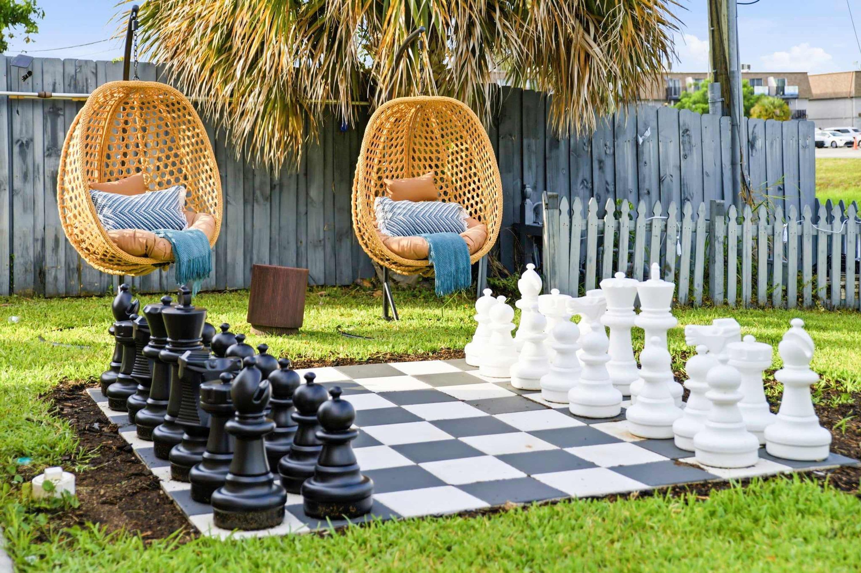 Un échiquier à carreaux avec des pièces d’échecs de deux couleurs, entouré d’une zone herbeuse où se trouvent deux chaises en osier et, au fond, une clôture en bois.