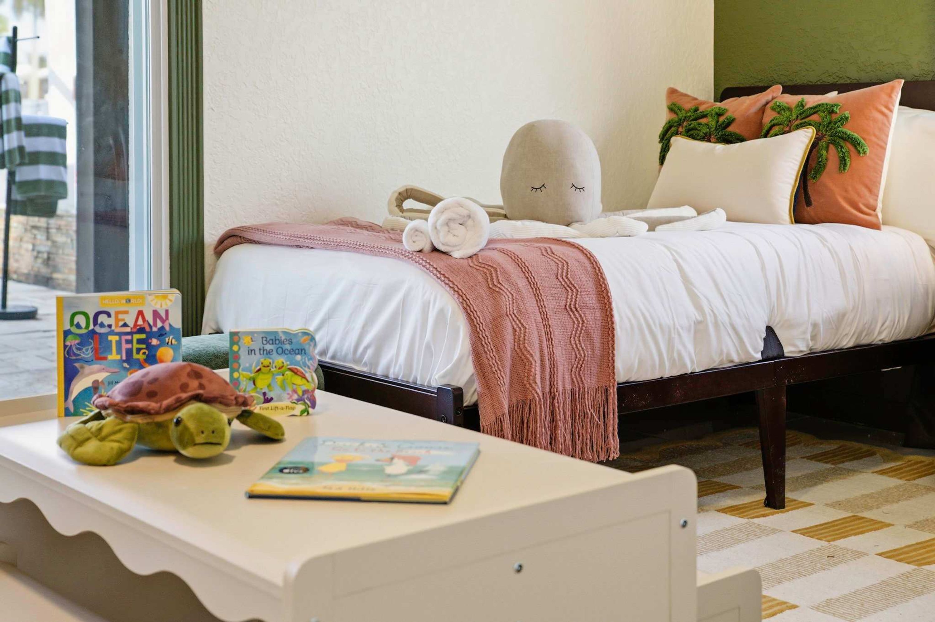 A neatly made bed with a white comforter, a pink knitted throw, and a pillow with a palm tree design.