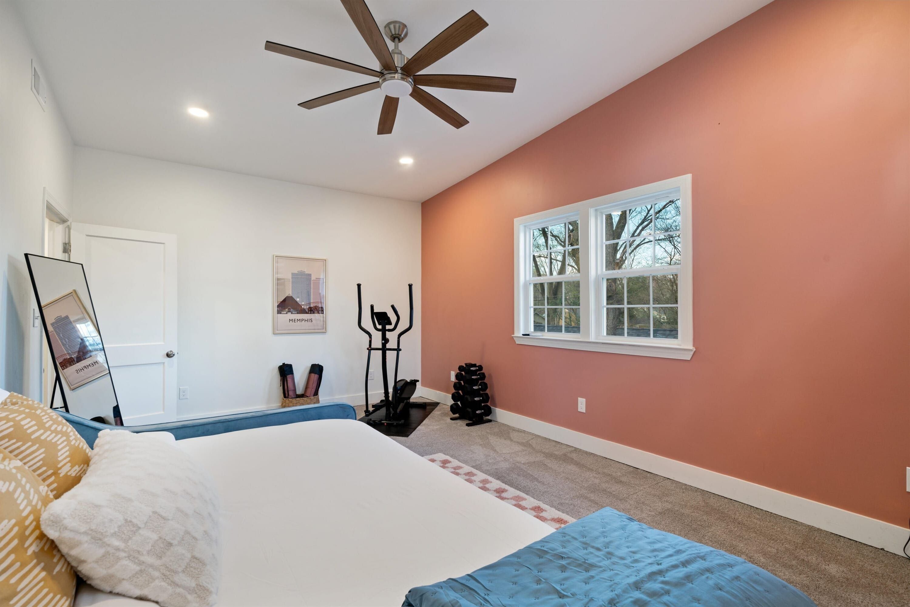 A bedroom with a bed, a ceiling fan, exercise equipment, and a window with a view of trees.