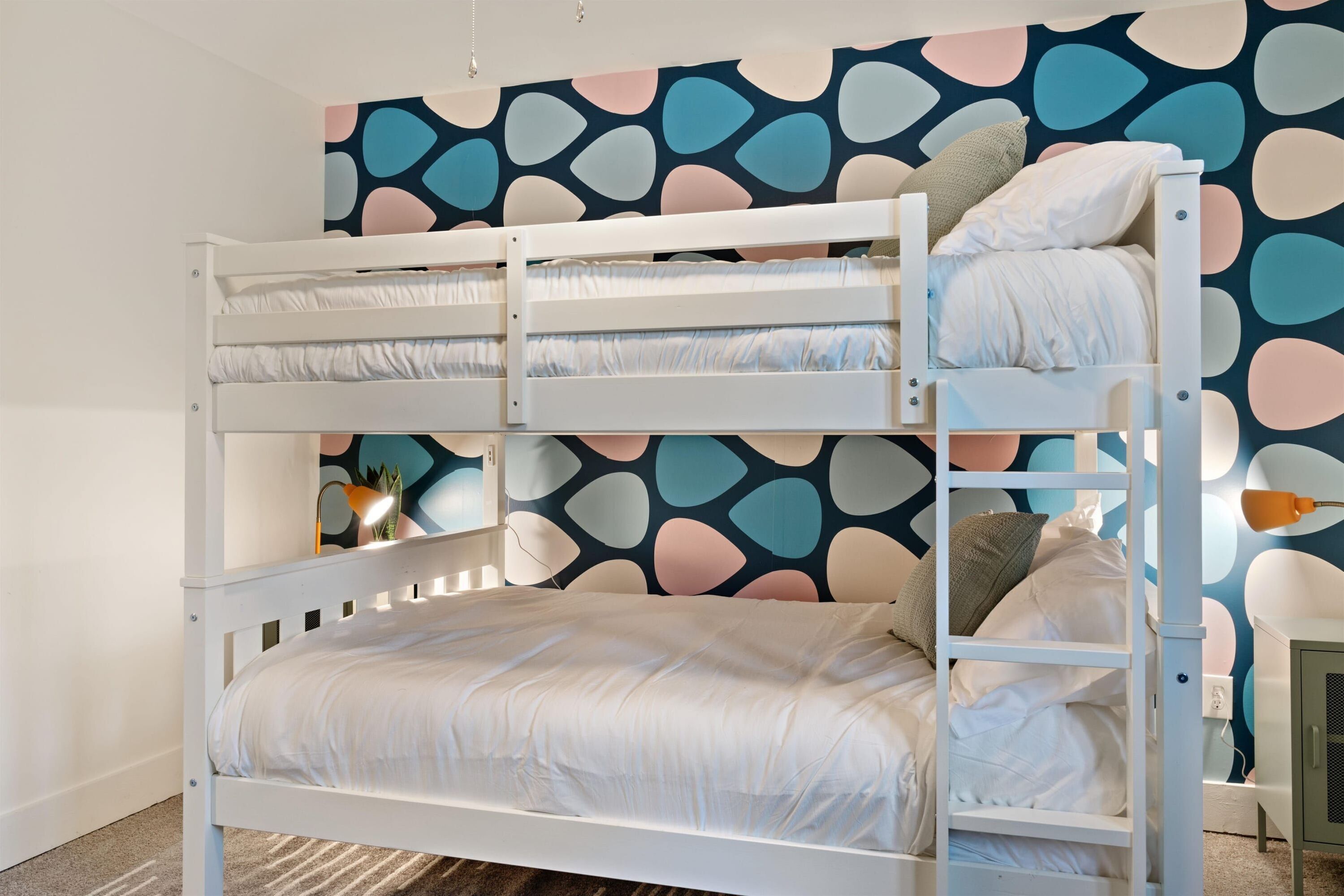 A bunk bed with a white frame and bedding, a wall with a colorful abstract pattern, and a small table with a lamp.