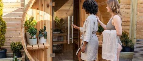 A wooden sauna with a glass door, surrounded by potted plants.