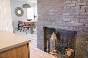 Uma cozinha com lareira de tijolos, uma área de jantar com cadeiras e mesa, e uma luminária pendente.