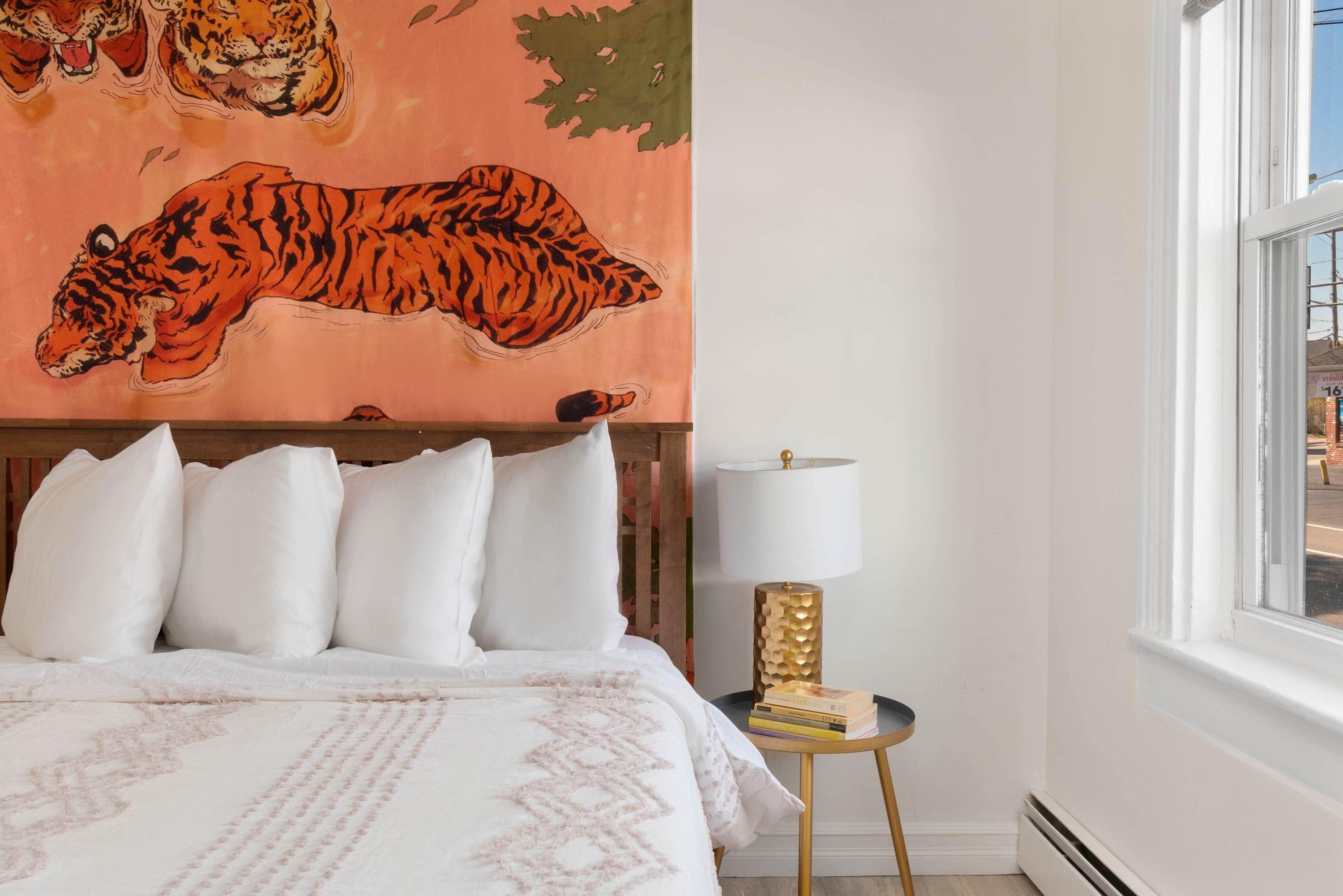 A bedroom with a bed, pillows, a wall tapestry of a tiger, a lamp, and a window.