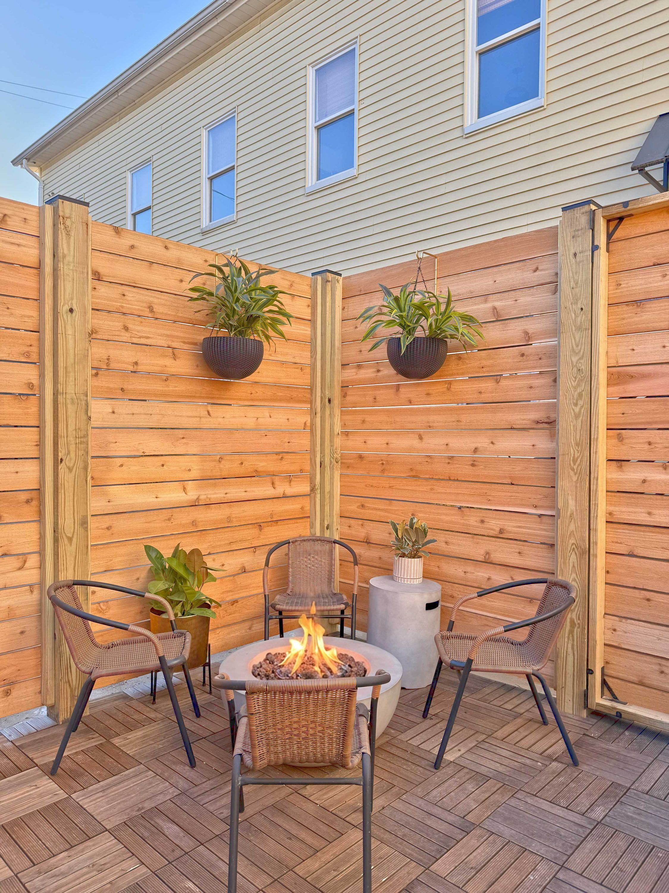 A patio area with a fire pit, two wicker chairs, and potted plants against a wooden fence.