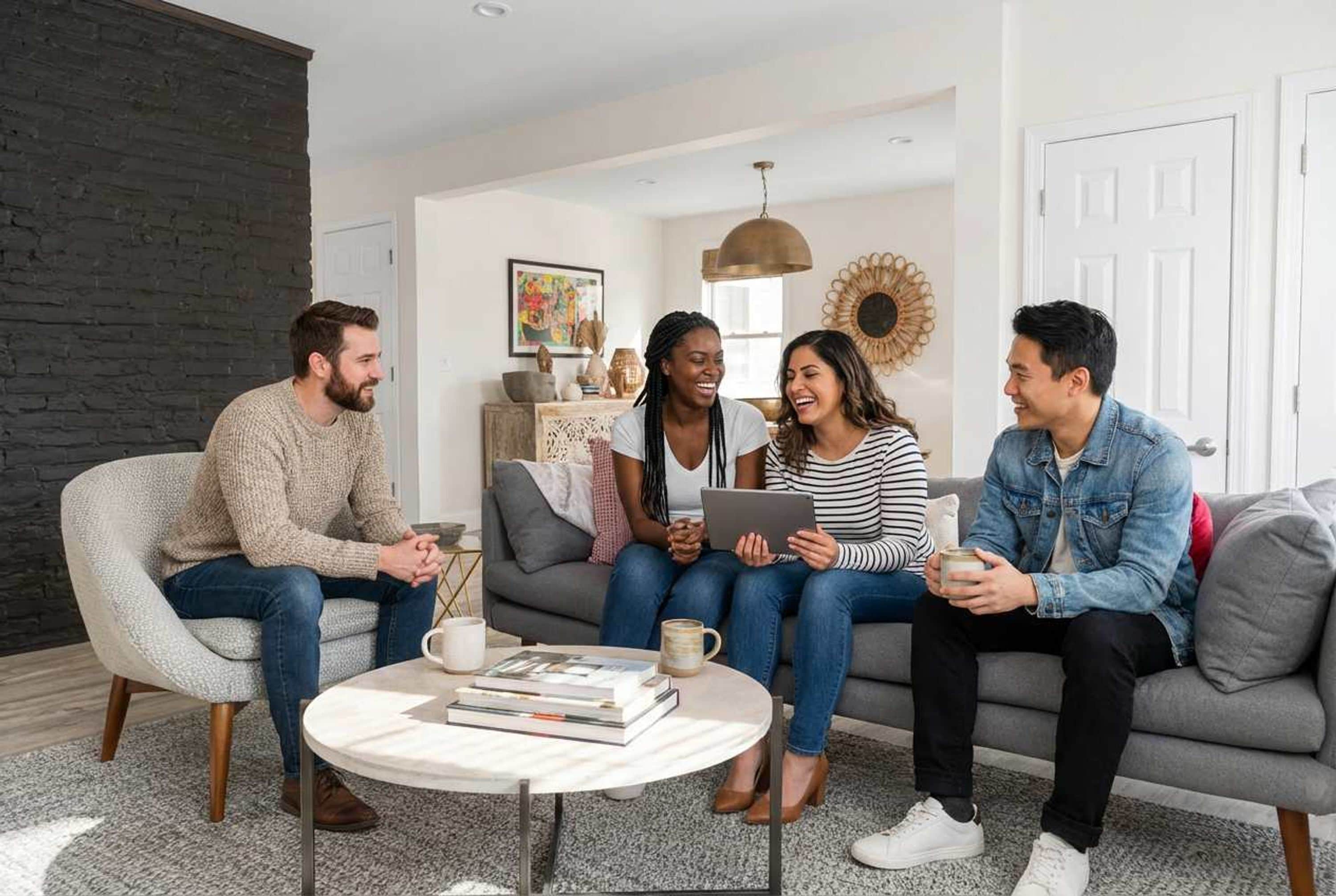A group of four people sitting on a grey sofa in a modern living room with a round coffee table, books, and a decorative wall piece.