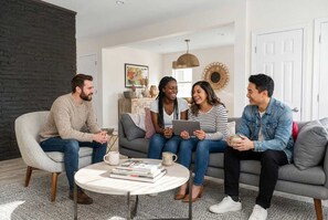 Un grupo de cuatro personas sentadas en un sofá gris en una sala de estar moderna, con una mesita redonda, libros y un adorno en la pared.