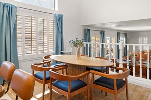 Un coin repas avec une table ronde et six chaises, un vase rempli de fleurs et une vue sur l’extérieur grâce à de grandes fenêtres ornées de rideaux bleus.