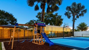 A playground with a slide and swings, a basketball court, and a wooden fence. - Resort-style Oasis Pool Mini Golf Playground (Largo)