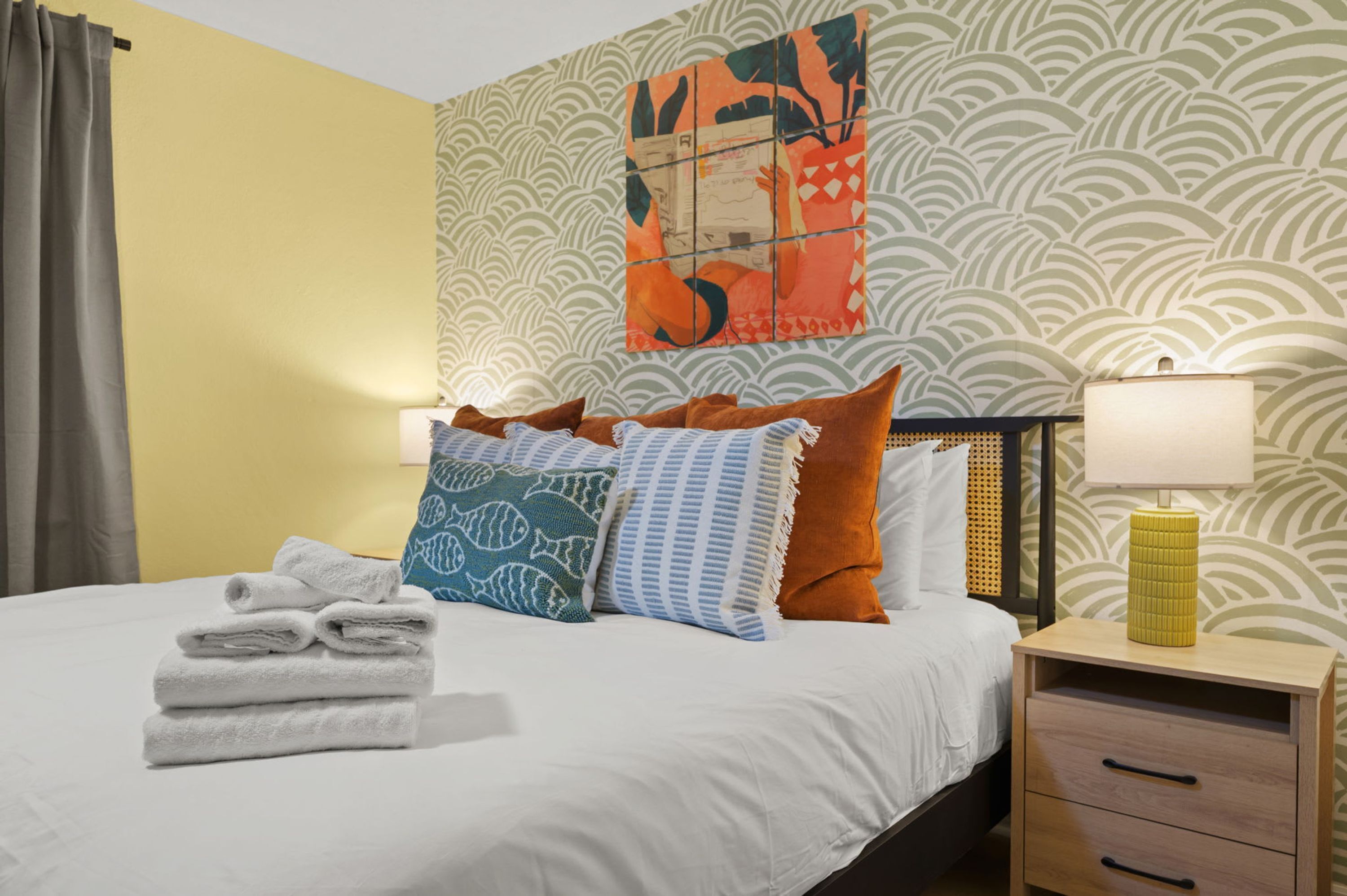 A hotel room with a bed, pillows, a nightstand, a lamp, and a wall with a patterned wallpaper.
