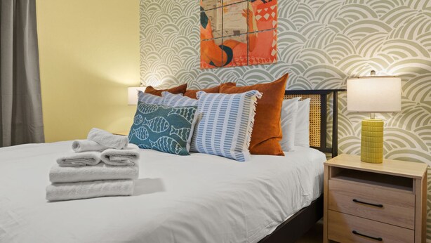 A hotel room with a bed, pillows, a nightstand, a lamp, and a wall with a patterned wallpaper. - Resort-style Oasis Pool Mini Golf Playground (Largo)