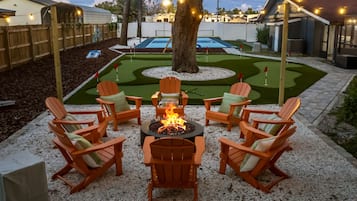 A backyard with a fire pit surrounded by Adirondack chairs, a small putting green, and a tree.