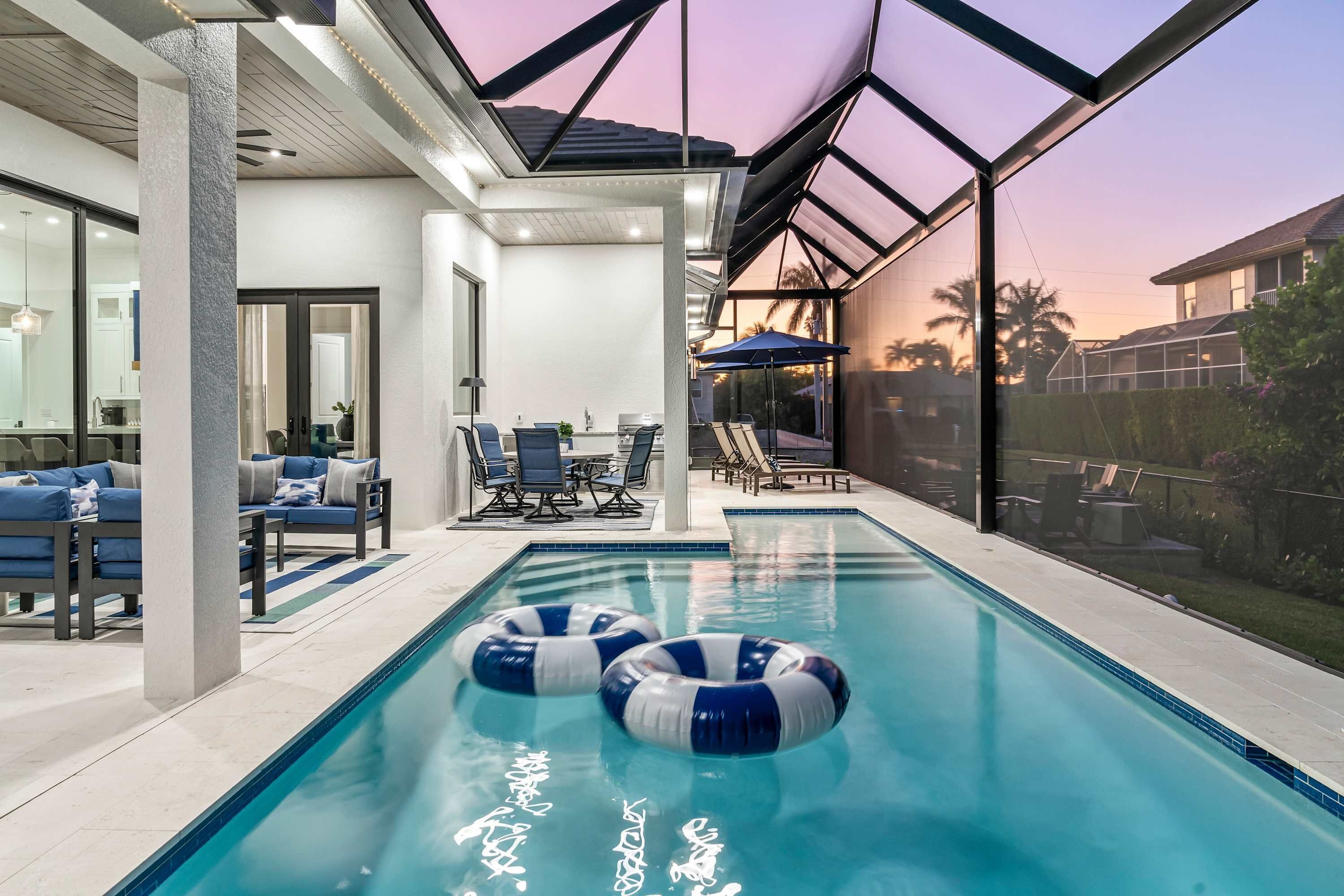An indoor swimming pool with two water slides, a seating area with chairs and a table, and a view of the sunset through a large window.