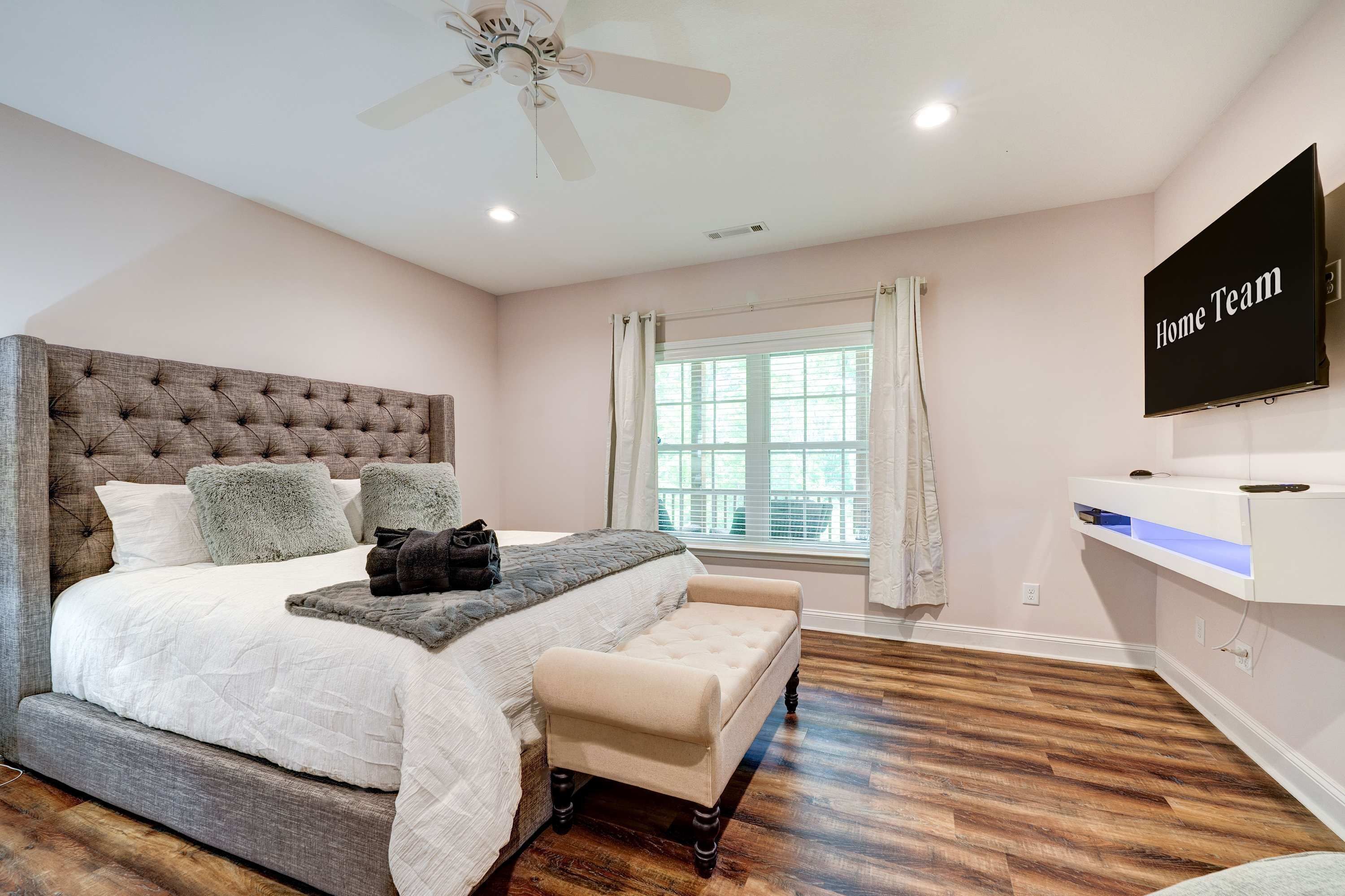 A bedroom with a large bed, a ceiling fan, a TV, and a window with curtains.