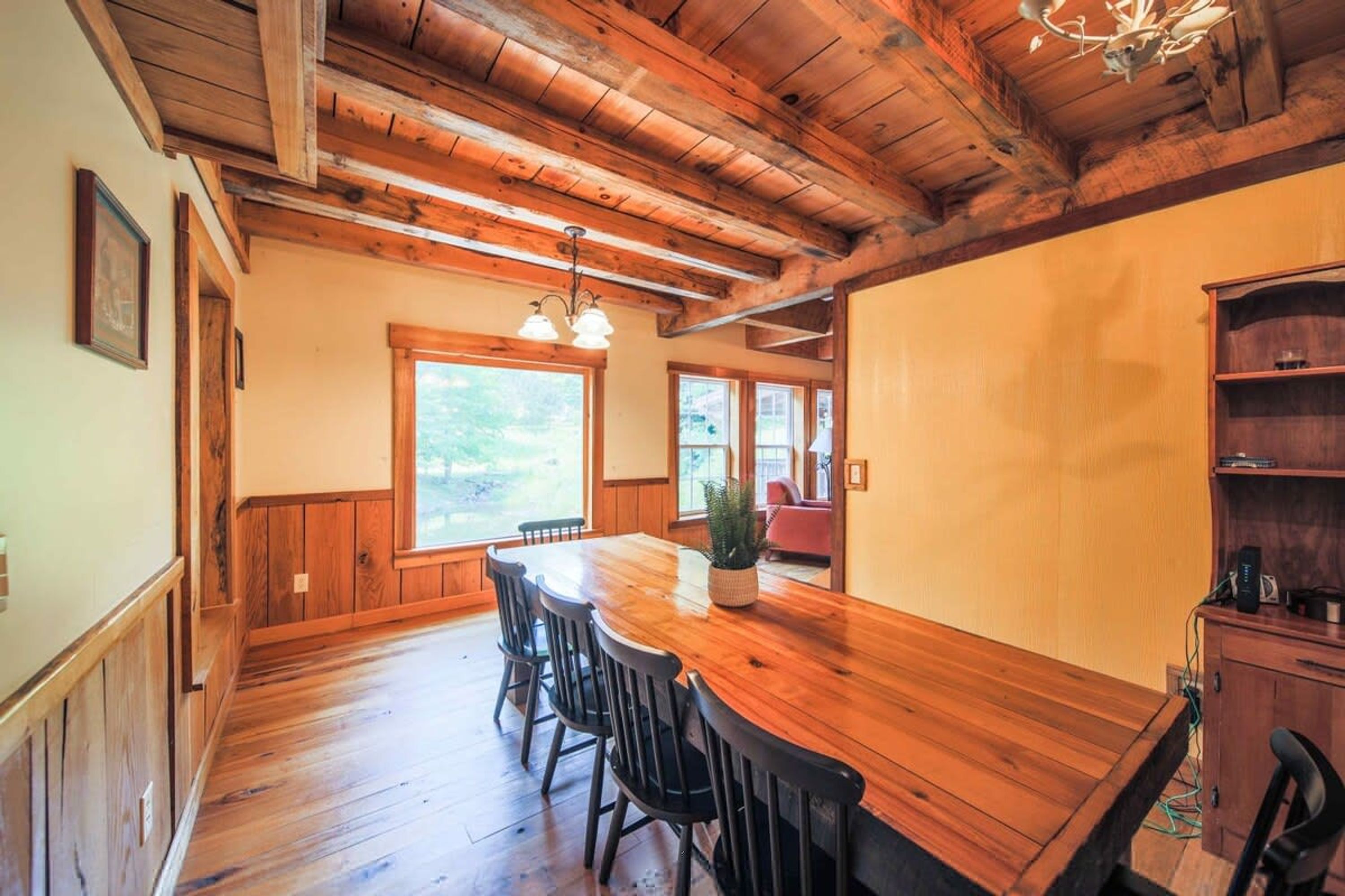 A wooden dining area with a long table and chairs, a chandelier, and a view of the outdoors through large windows.