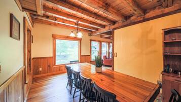 A wooden dining area with a long table and chairs, a chandelier, and a view of the outdoors through large windows.