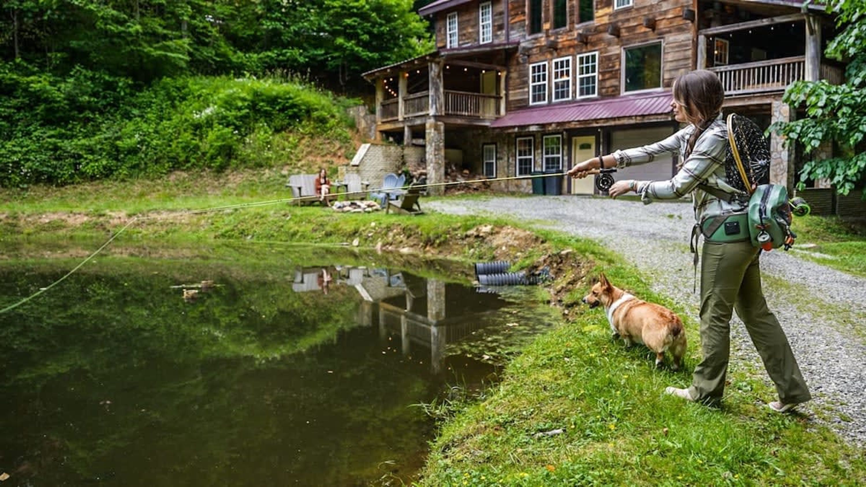 A person fishing by a pond with a dog, a wooden building in the background, and a clear sky.
