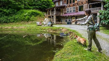 A person fishing by a pond with a dog, a wooden building in the background, and a clear sky.