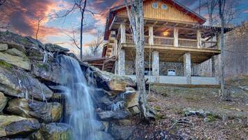 A wooden cabin with a stone foundation, a waterfall in front, and a sunset in the background.