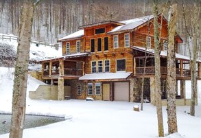 A multi-story wooden cabin with a balcony, a garage, and a snow-covered ground.