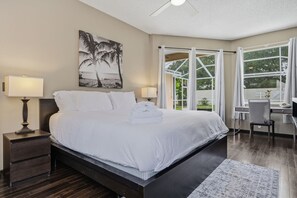 A hotel room with a large bed, two bedside tables, a desk, and a chair. There is a window with curtains and a view of greenery outside. - Heated Pool Game Room Outdoor Fun Sleeps 6 (Tampa)