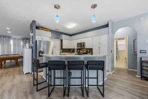A modern kitchen with a central island, bar stools, and a pool table in the adjacent room. - Heated Pool Game Room Outdoor Fun Sleeps 6 (Tampa)