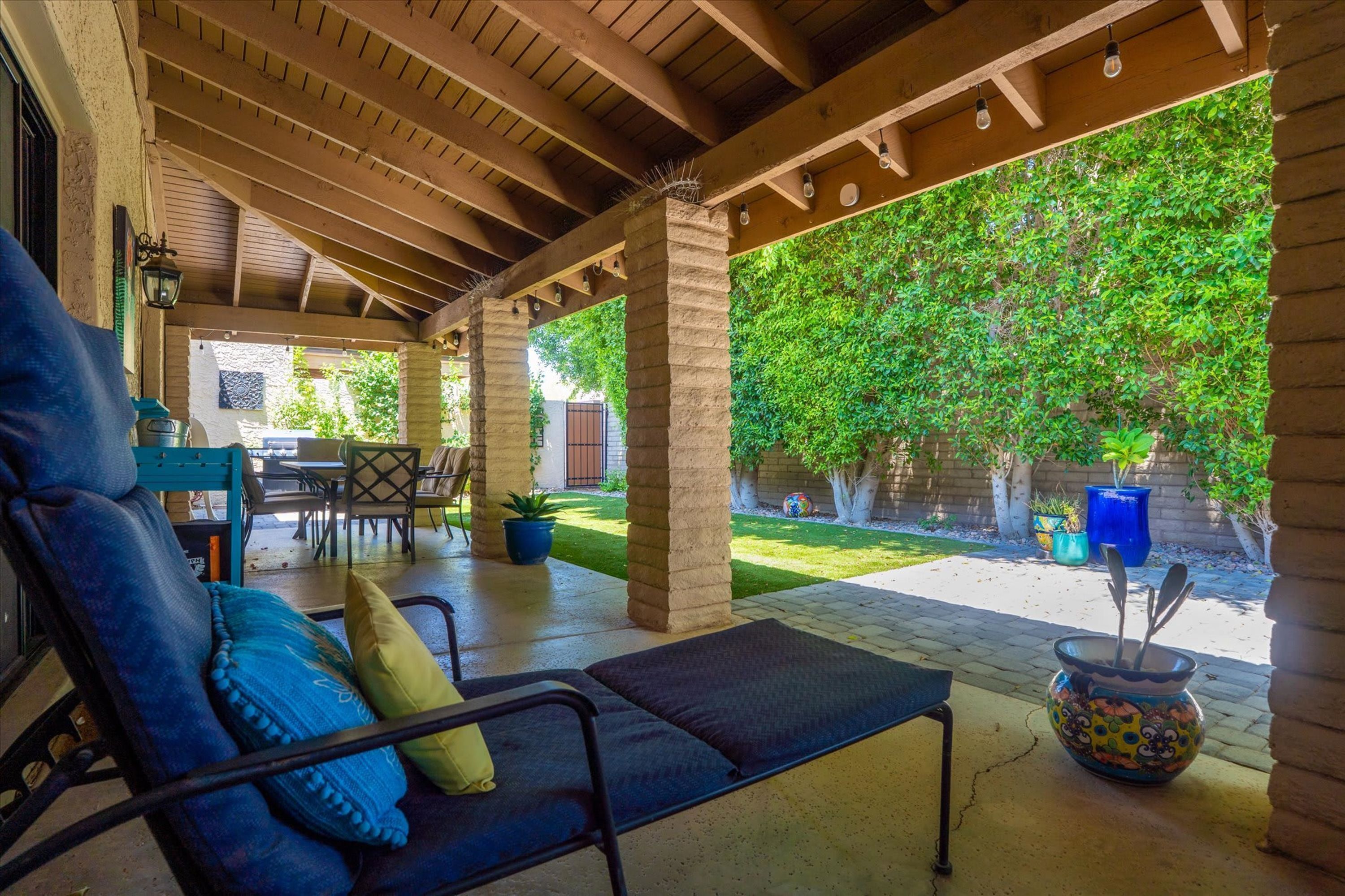 A covered patio with a lounge chair, a table, and chairs, surrounded by greenery and a brick wall.