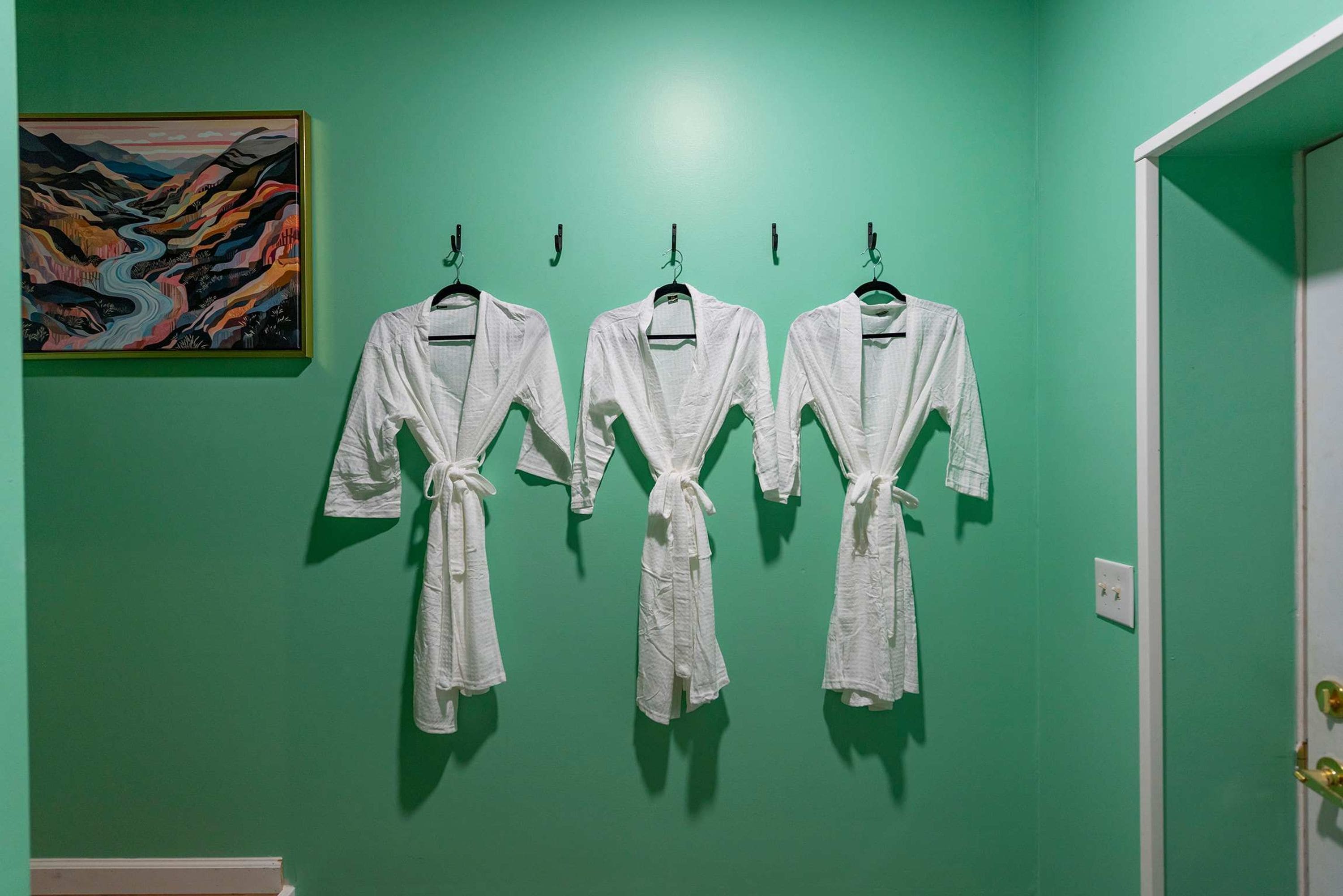 Three white bathrobes hanging on hooks against a green wall, with a painting above them.