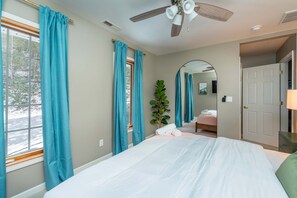 Une chambre à coucher avec un lit, un ventilateur de plafond, une fenêtre avec des rideaux, un miroir et une porte.