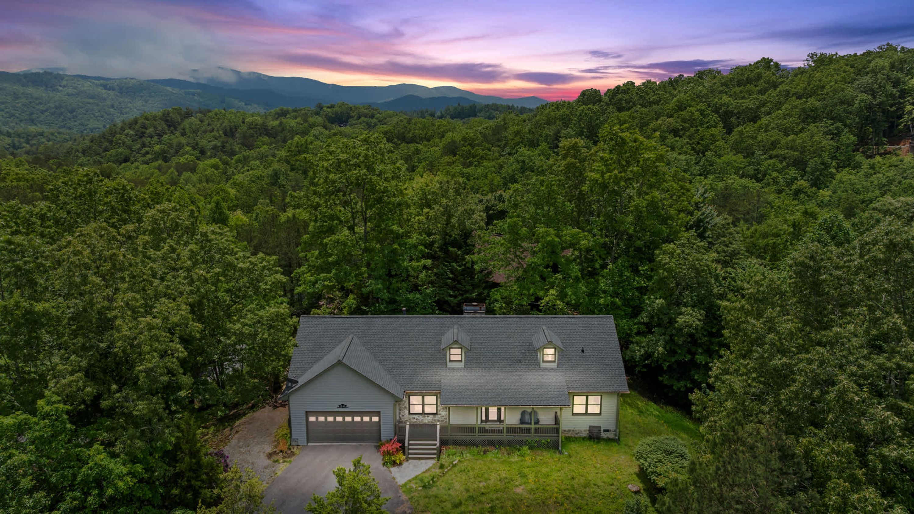 Une maison à deux étages avec un garage, entourée d’une forêt dense, et une chaîne de montagnes en arrière-plan.