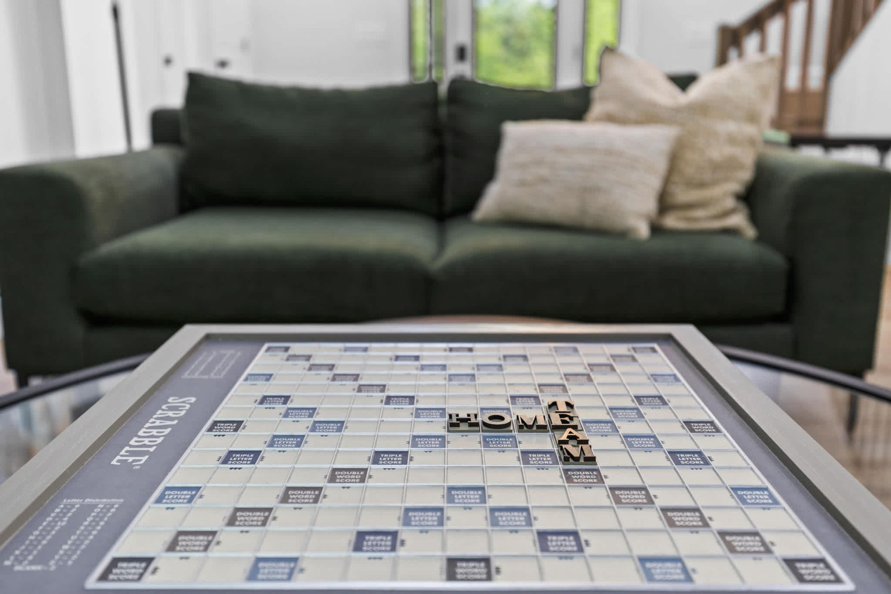A Scrabble board game with the word 'HOME' on it, placed on a glass table in front of a green sofa with two pillows.