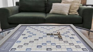 Et Scrabble-spil med ordet 'HOME' på, placeret på et glasbord foran en grøn sofa med to puder.