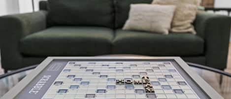 Un jeu de société Scrabble, avec le mot « HOME » inscrit sur le plateau, est posé sur une table en verre devant un canapé vert agrémenté de deux coussins.