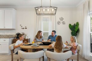 Eine Familie speist in einer modernen Küche mit weißen Schränken, einer zentralen Kochinsel und einem großen Fenster.