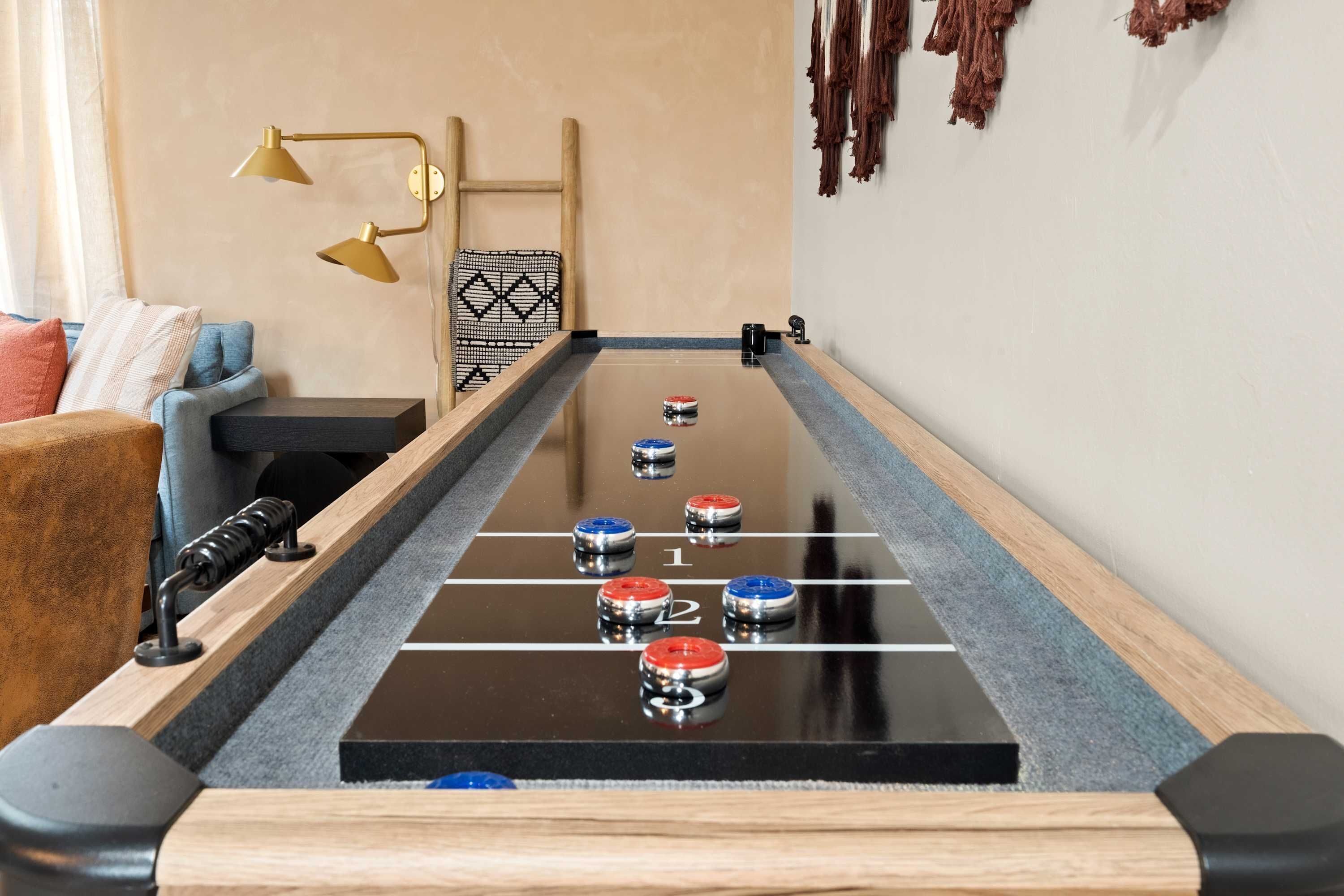 A shuffleboard table with pucks in various colors and numbers, a sofa, a lamp, and a wall-mounted decorative item.