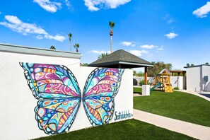 A building with a colorful butterfly mural on the wall, a small play structure, and a clear blue sky. - Sonoran Grove Epic Yard With Two Pickleball Courts (Scottsdale)
