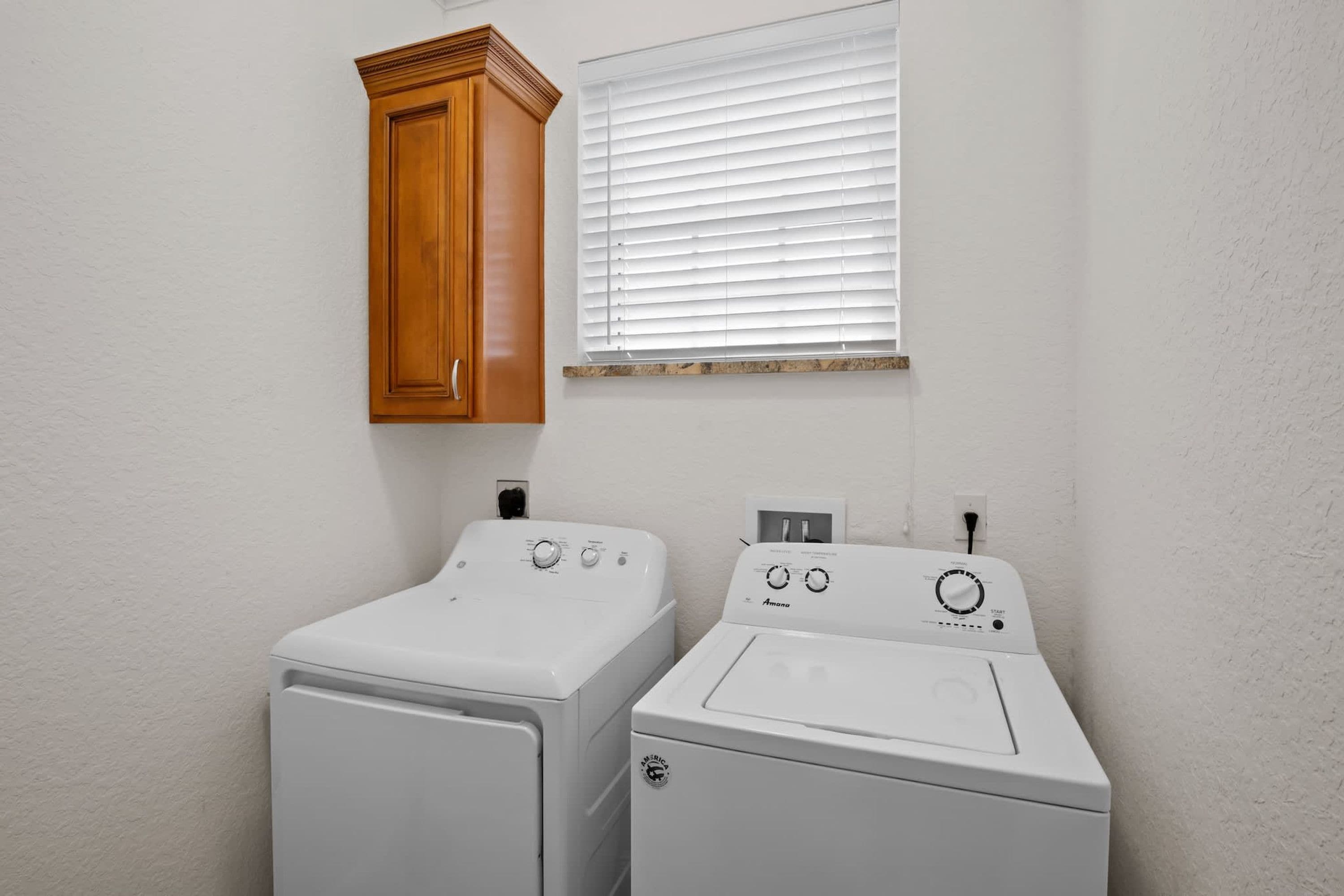 A laundry room with two white washing machines, a wooden cabinet, and a window with blinds.