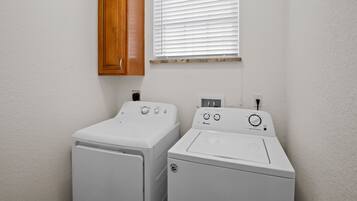 A laundry room with two white washing machines, a wooden cabinet, and a window with blinds.