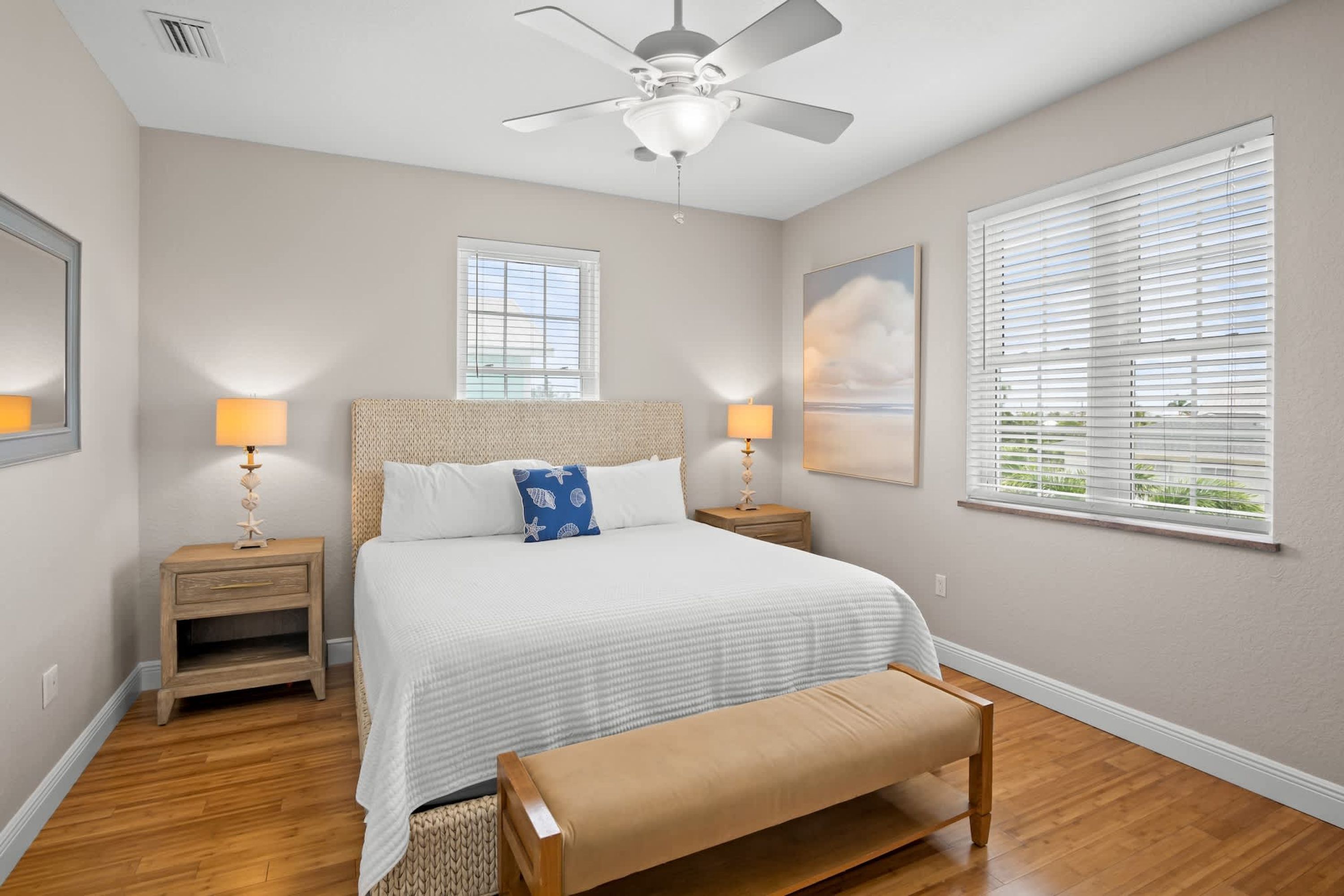 A bedroom with a bed, two bedside tables, a bench, a ceiling fan, and two windows with blinds.