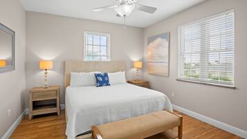 A bedroom with a bed, two bedside tables, a bench, a ceiling fan, and two windows with blinds.