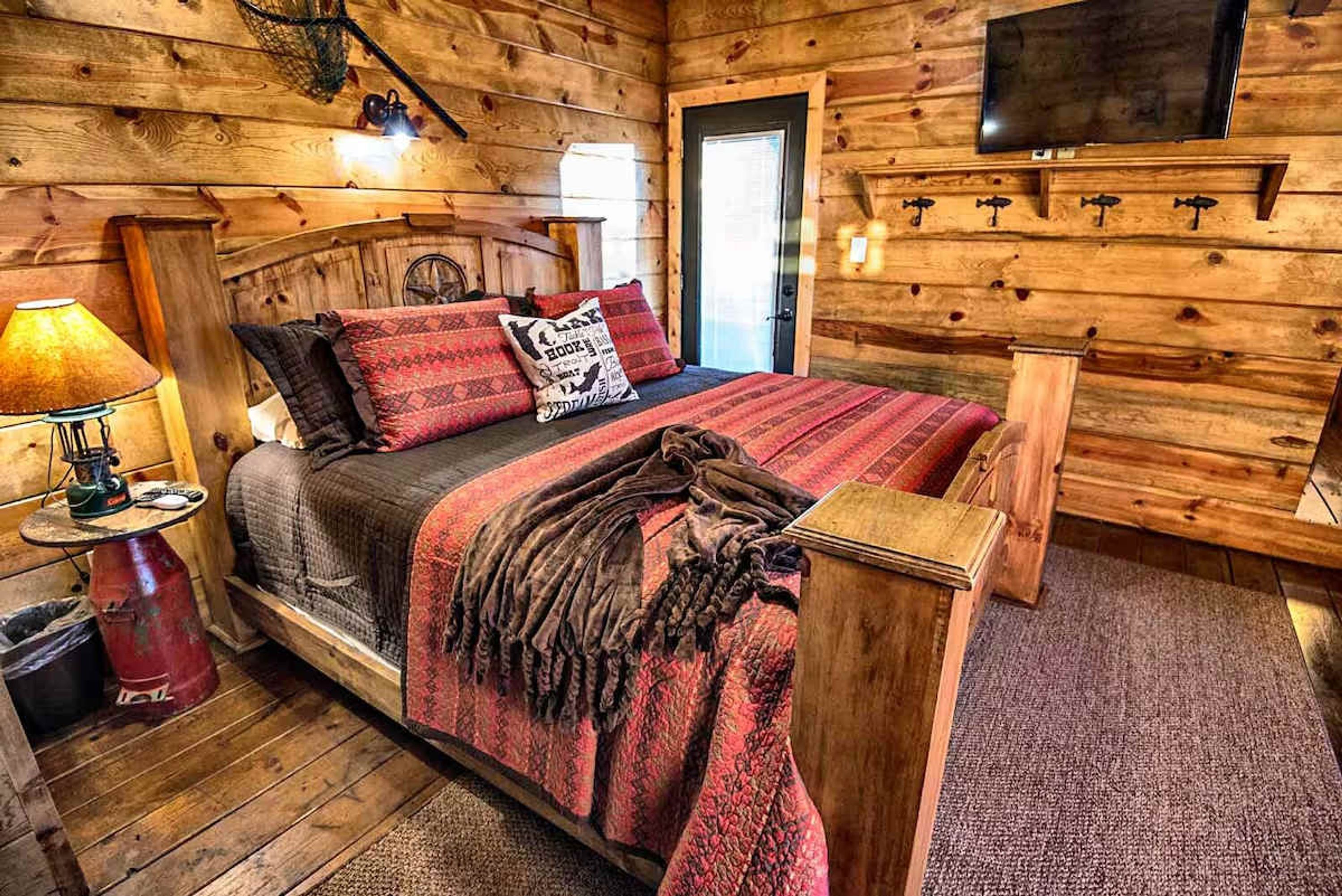 Interior de una cabaña de madera con una cama, un televisor montado en la pared y una lámpara sobre una mesita.