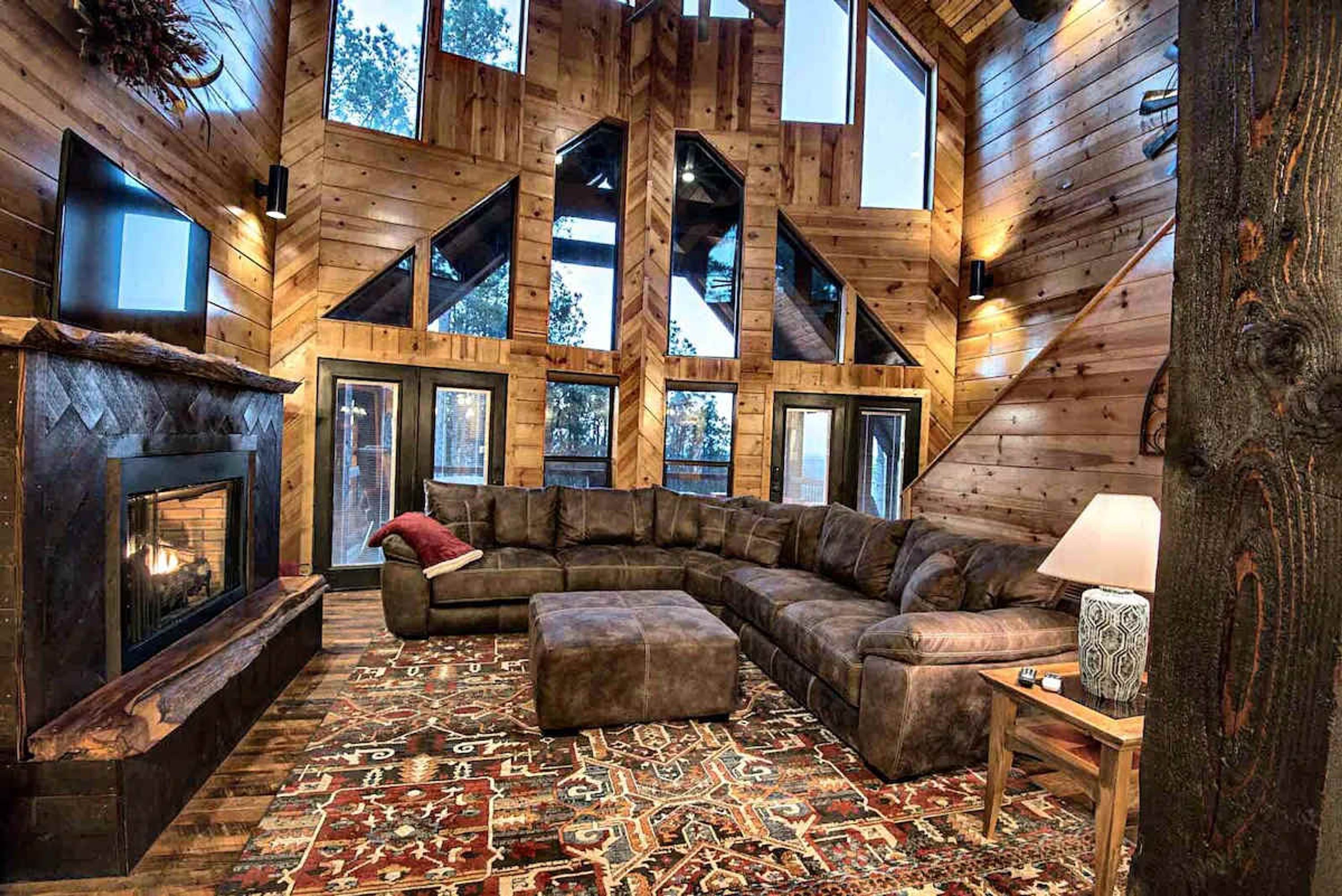 A log cabin interior with a large L-shaped sectional sofa, a fireplace, and a patterned rug.