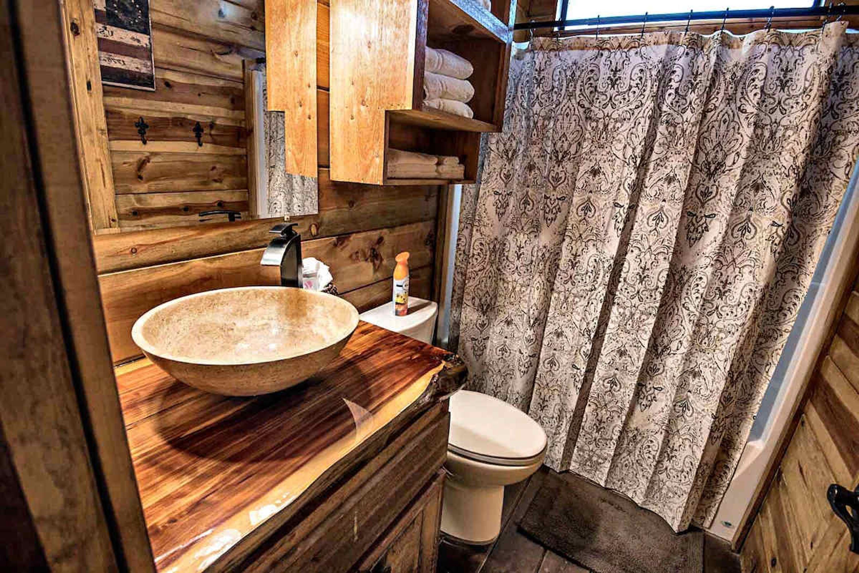 A rustic bathroom with a wooden vanity, a large bowl sink, a toilet, and a patterned shower curtain.